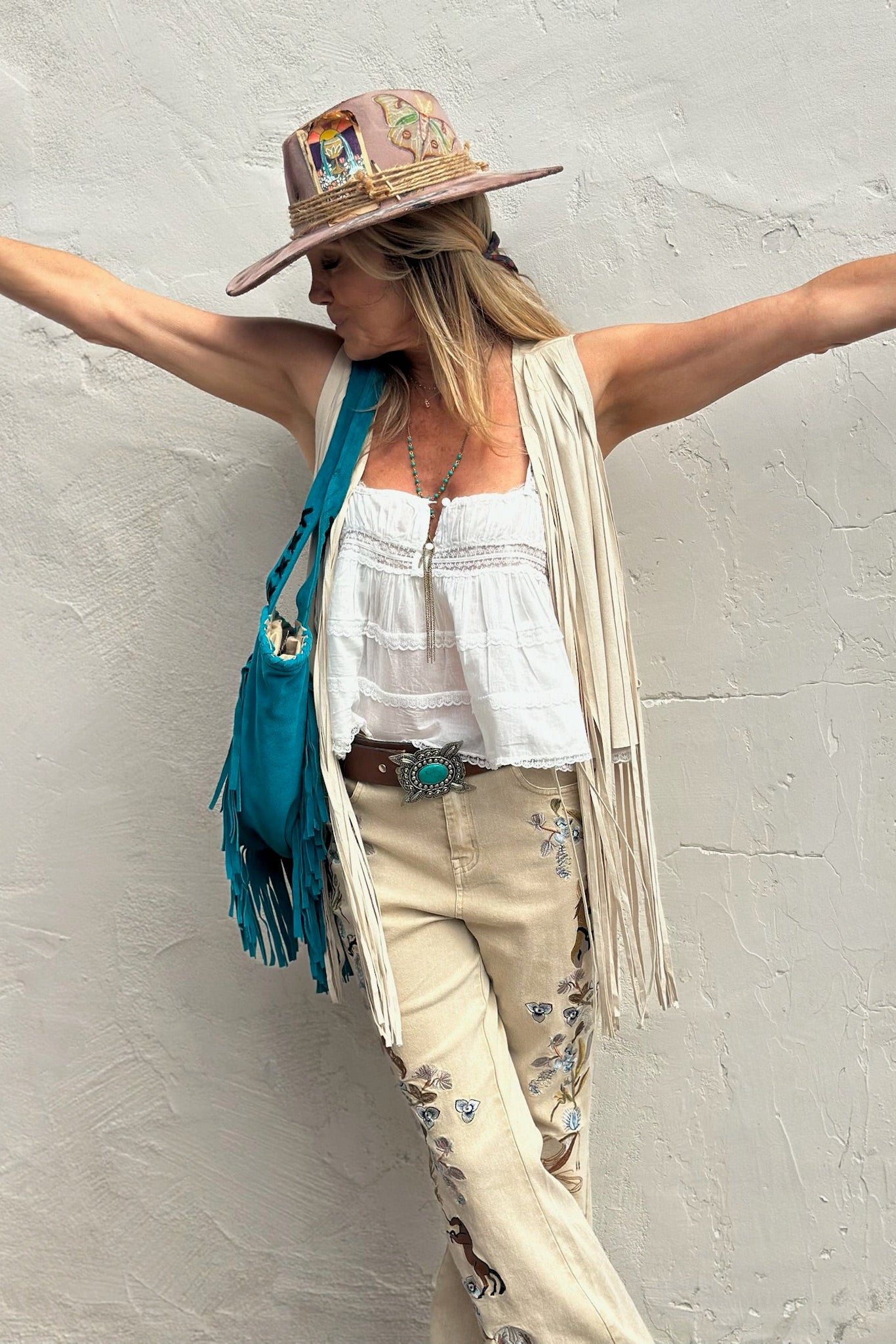 Woman in a white top, beige pants, and colorful hat with arms outstretched against a gray wall.
